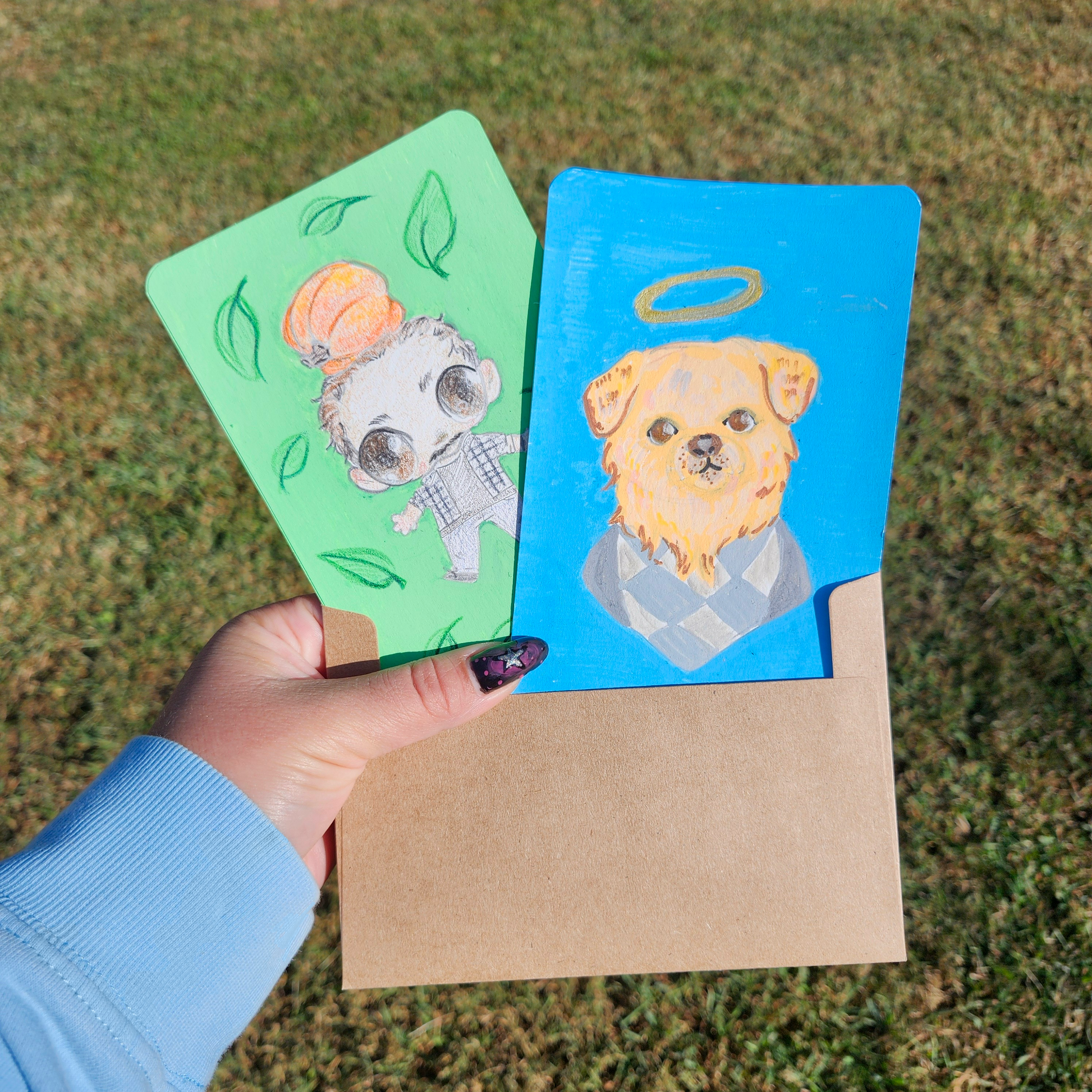 Hand holding 2 illustrations, one of a man with a pumpkin, the other of a dog with a halo, in front of a grassy background.